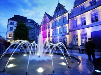 Beleuchteter Brunnen vor historischen Gebäuden in Abenddämmerung, Personen passieren.