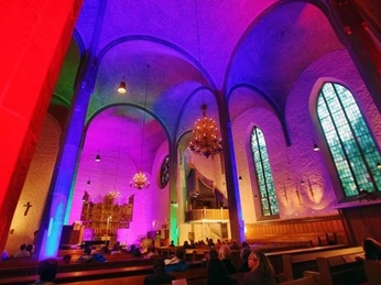 Kircheninnenraum mit leuchtenden, bunten Lichteffekten und Besuchern auf den Holzbänken.