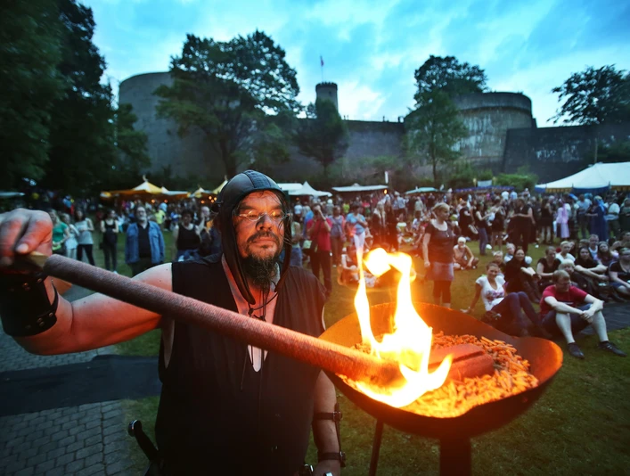 Ein mittelalterlich gekleideter Mann entzündet ein Feuer vor einer Menschenmenge nahe einer Burg bei Dämmerung.