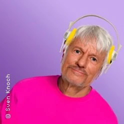 Ein älterer Mann mit weißen Haaren und gelbem Kopfhörer trägt ein pinkes Oberteil vor lila Hintergrund.