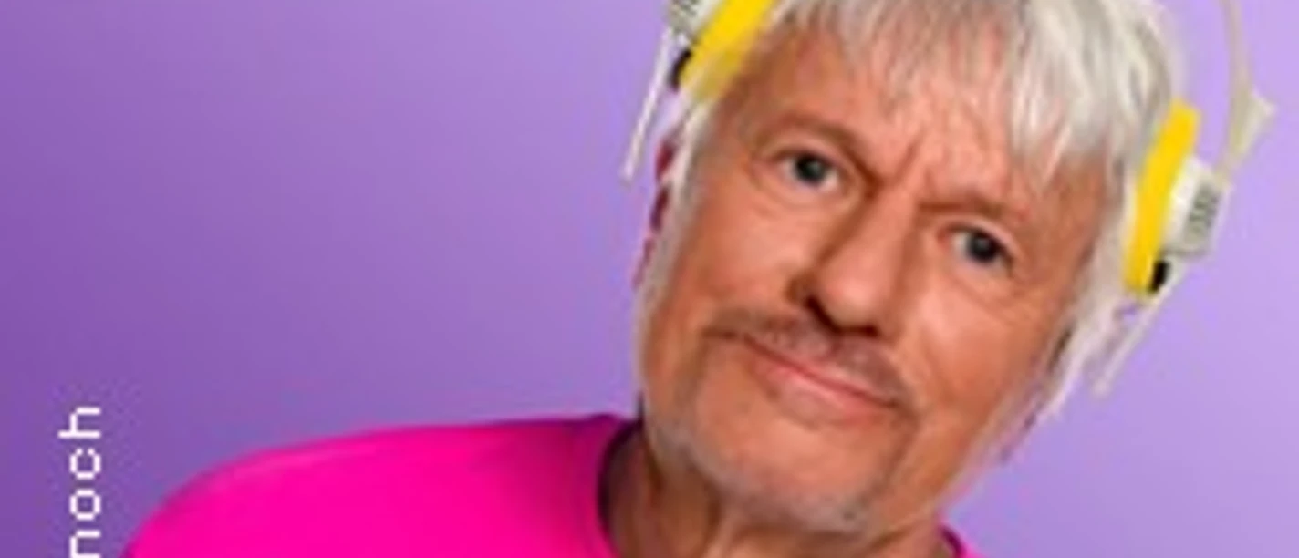 Ein älterer Mann mit weißen Haaren und gelbem Kopfhörer trägt ein pinkes Oberteil vor lila Hintergrund.