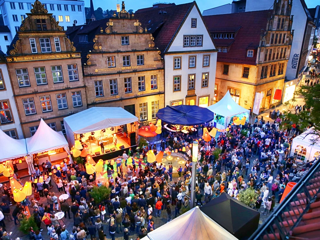 Leinewebermarkt ©Bielefeld Marketing Menschenmenge feiert auf einem lebhaften Straßenfest, umgeben von historischen Fachwerkhäusern bei Abenddämmerung.
