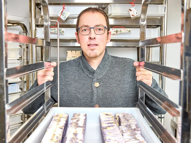 Seifenmanufaktur Würfel Person steht zwischen Metallregalen mit handgefertigten Seifenstücken in einer Manufaktur.