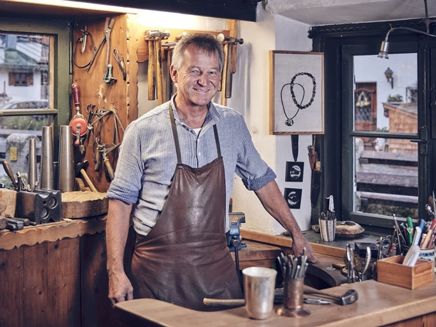 Silberschmiede Adam Ein Silberschmied steht lächelnd an seinem Arbeitsplatz mit Werkzeugen an der Wand.