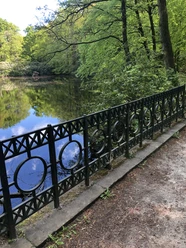 Rundteichbrücke Ein ruhiger, runder Teich von Bäumen umgeben, mit schmiedeeisernem Geländer im Vordergrund.