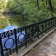 Rundteichbrücke Ein ruhiger, runder Teich von Bäumen umgeben, mit schmiedeeisernem Geländer im Vordergrund.