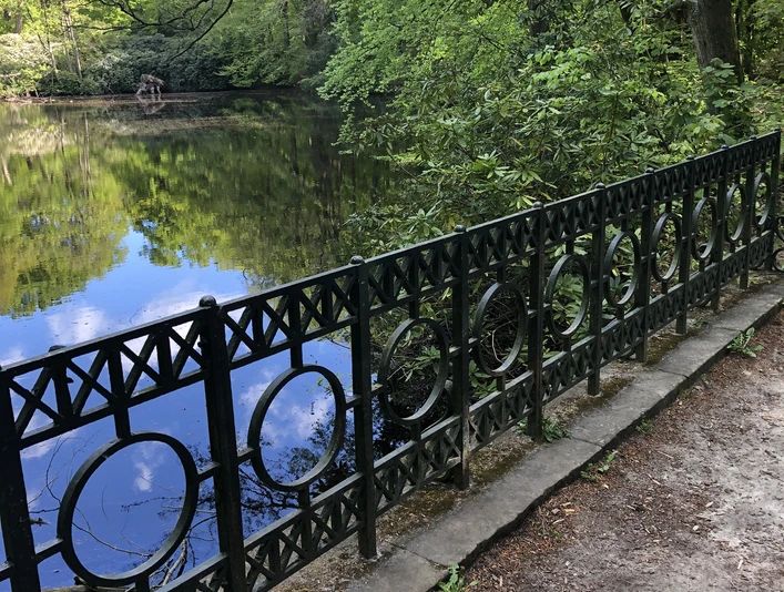 Rundteichbrücke Ein ruhiger, runder Teich von Bäumen umgeben, mit schmiedeeisernem Geländer im Vordergrund.