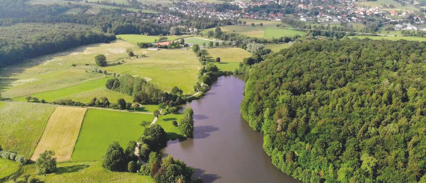 Nethestausee-Neuenheerse Luftaufnahme des Nethestausees, umgeben von dichten Wäldern und Feldern, Dorf im Hintergrund.