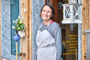 Ungnadner Keramik Eine Frau in Schürze steht lächelnd vor einem Keramikgeschäft mit Blumenarrangement.
