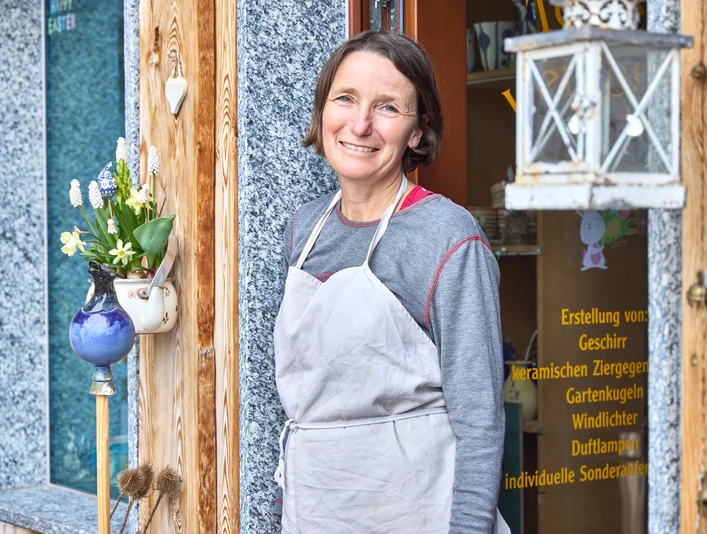 Ungnadner Keramik Eine Frau in Schürze steht lächelnd vor einem Keramikgeschäft mit Blumenarrangement.
