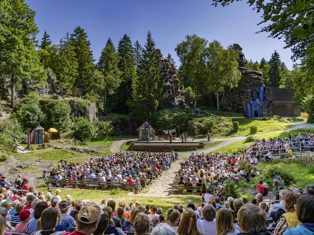 Hotzenplotz-Kulisse-2025_Foto_Dirk_Rückschloss.jpg Ein Freilufttheater mit Zuschauern vor einer Waldbühne, umgeben von Felsen und Bäumen.