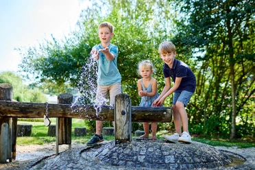Hinterhermsdorf Freizeitanlage Drei Kinder spielen fröhlich an einem Wasserspielplatz, spritzen Wasser und lachen, umgeben von grünen Bäumen und sonnigem Wetter.