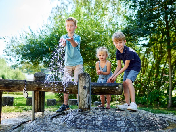 Hinterhermsdorf Freizeitanlage Drei Kinder spielen fröhlich an einem Wasserspielplatz, spritzen Wasser und lachen, umgeben von grünen Bäumen und sonnigem Wetter.