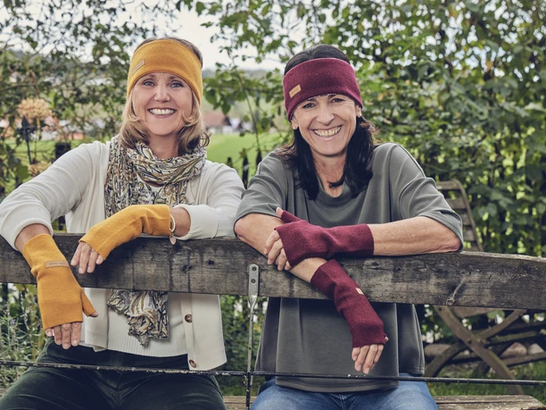 Wuide Henna Zwei lächelnde Frauen mit Stirnbändern und Armstulpen ruhen auf einem Holzzaun.