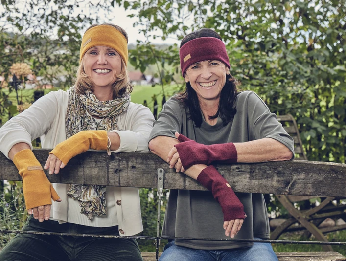 Wuide Henna Zwei lächelnde Frauen mit Stirnbändern und Armstulpen ruhen auf einem Holzzaun.