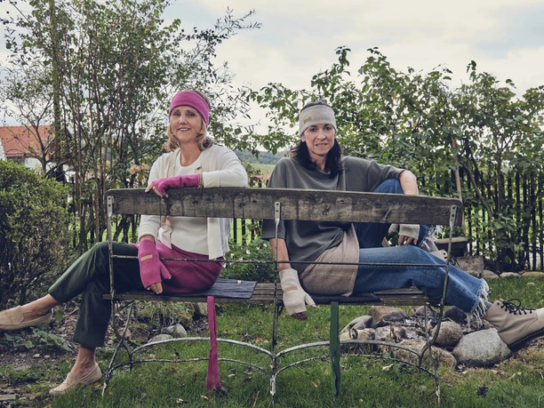 Wuide Henna Zwei Frauen sitzen auf einer Parkbank, tragen farbenfrohe Mützen und Handschuhe.