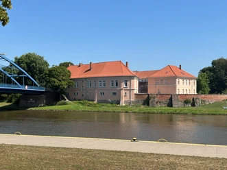 Castle complex with red roof on the banks of the Weser, surrounded by trees, under a clear blue sky.