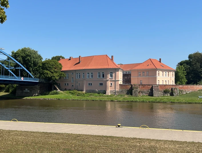 Schlossanlage mit rotem Dach am Weserufer, umgeben von Bäumen, bei klarem blauem Himmel.Castle complex with red roof on the banks of the Weser, surrounded by trees, under a clear blue sky.Slotskompleks med rødt tag på bredden af Weser, omgivet af træer, under en klar blå himmel.Kasteelcomplex met rood dak aan de oever van de Weser, omringd door bomen, onder een strakblauwe hemel.