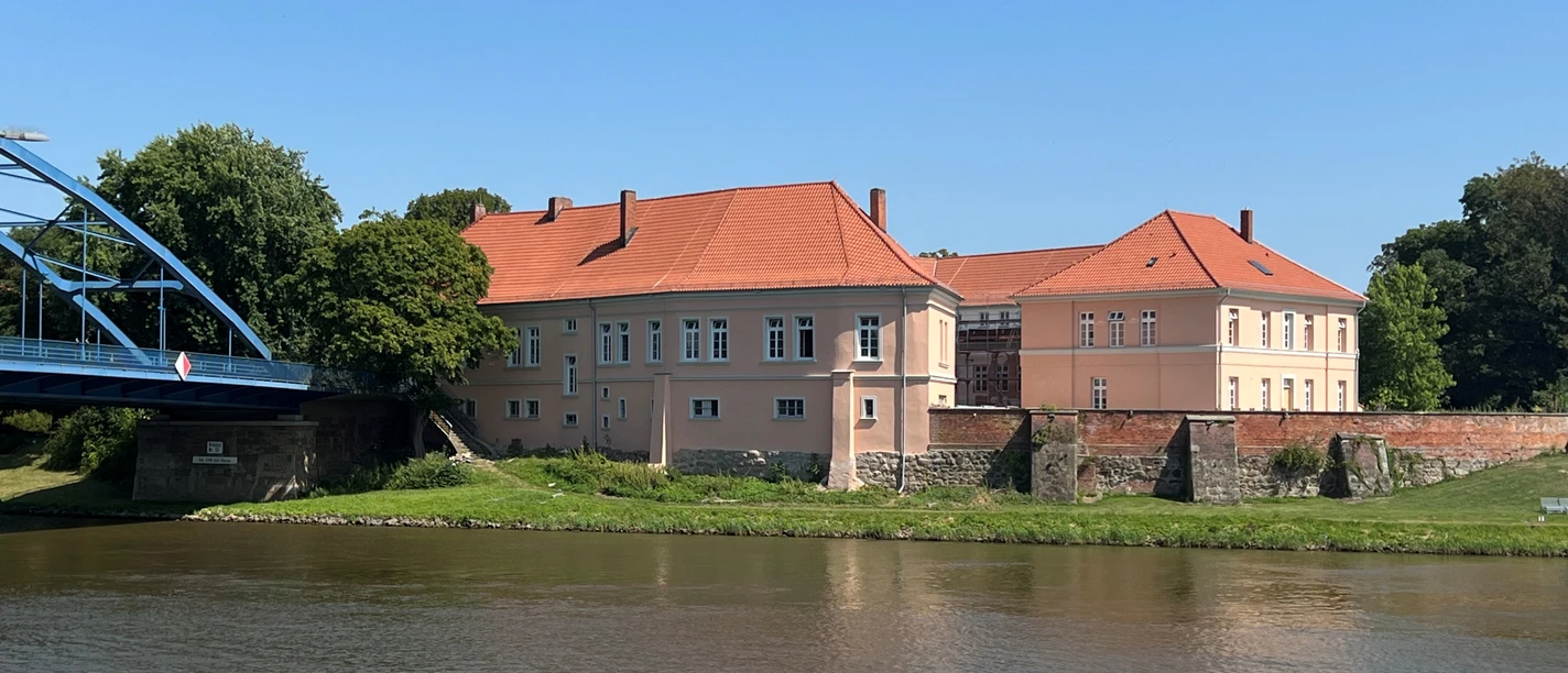 Grafenschloss Hoya Castle complex with red roof on the banks of the Weser, surrounded by trees, under a clear blue sky.