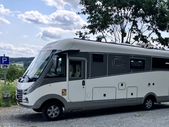Wohnmobilstellplatz_Stadt-Borgholzhausen (2).JPG Weiß-graues Wohnmobil auf Schotterparkplatz, umgeben von grünen Bäumen und hügeliger Landschaft.