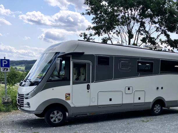 Wohnmobilstellplatz_Stadt-Borgholzhausen (2).JPG Weiß-graues Wohnmobil auf Schotterparkplatz, umgeben von grünen Bäumen und hügeliger Landschaft.