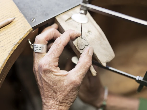 Susi´s Schmuckwerkstatt Eine Hand bearbeitet ein Herz-Motiv auf Holz in einer Werkstatt.