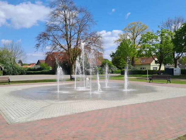 Brunnen Kögel-Willms-Platz Das Bild zeigt einen lebendigen Brunnen mit mehreren sprudelnden Wasserfontänen, umgeben von einer parkähnlichen Grünanlage und einem klaren blauen Himmel im Hintergrund.