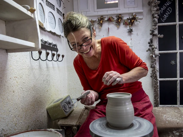 Töpferei Winsi Eine lächelnde Frau an der Töpferscheibe formt ein Gefäß in einer Werkstatt.