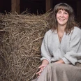 Studio. Birgit Sailer Eine lächelnde Frau sitzt vor einem Strohbündel in rustikaler Umgebung.