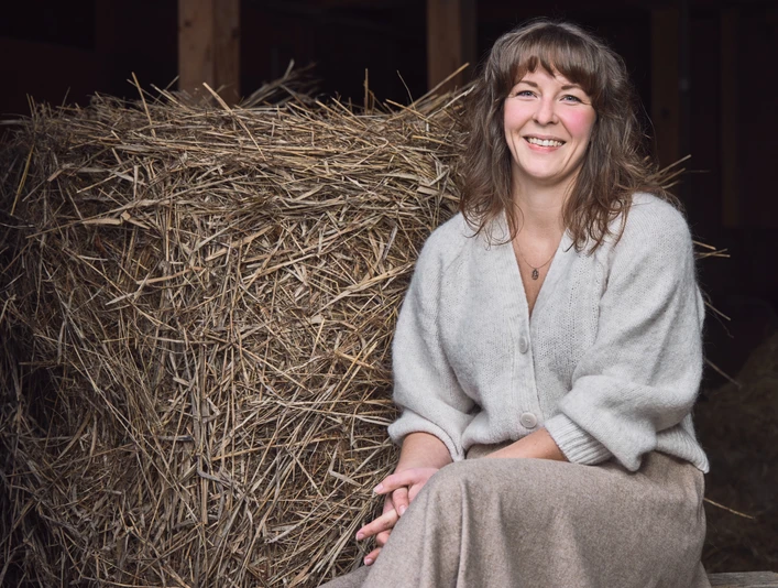 Studio. Birgit Sailer Eine lächelnde Frau sitzt vor einem Strohbündel in rustikaler Umgebung.