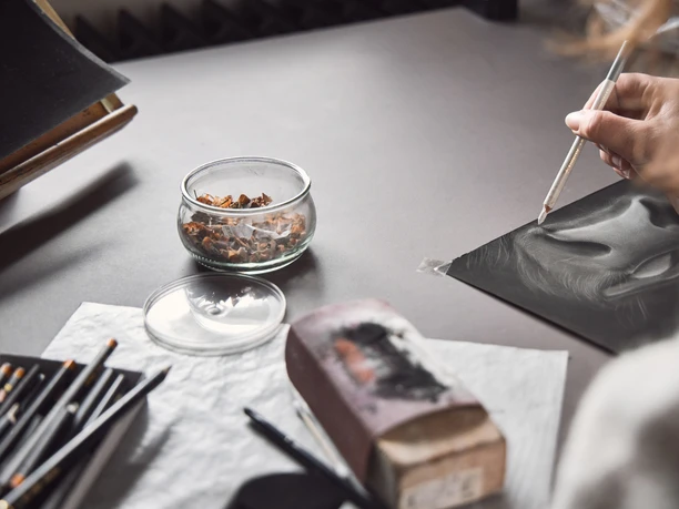 Studio. Birgit Sailer Eine Hand zeichnet mit einem weißen Buntstift ein Gesicht auf schwarzem Papier.