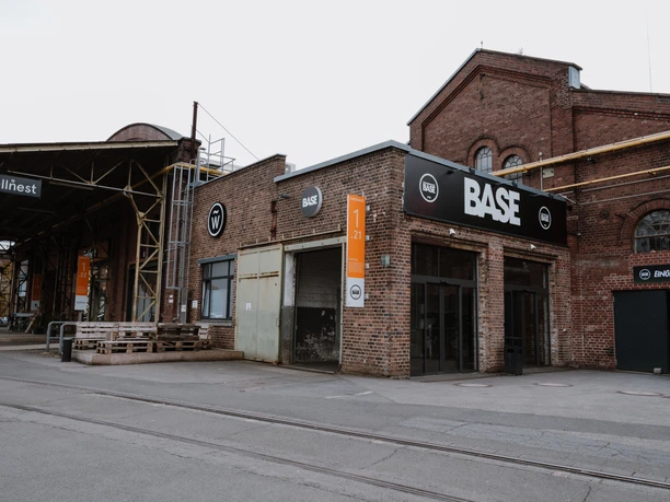 BASE draußen Das Bild zeigt die rote Backsteinfassade eines historischen Industriegebäudes mit einem großen BASE-Logo darüber. Daneben befinden sich Schilder mit der Hausnummer 1/21 und einem weiteren, kleineren Logo einer Coilfeder. Der Eingangsbereich ist modern gestaltet.