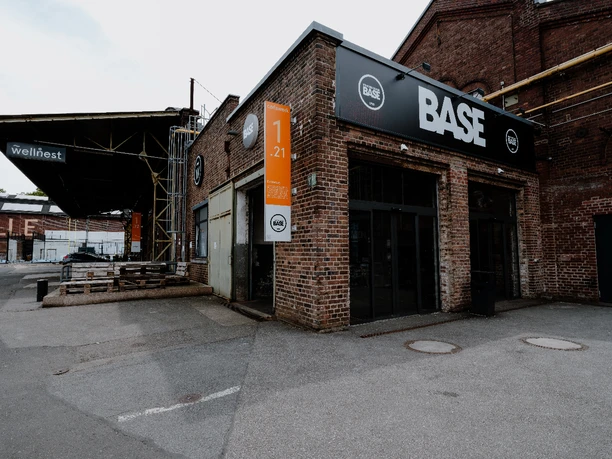 BASE draußen Das Bild zeigt einen modernen, aus roten Backsteinen errichteten Gebäudekomplex mit der Aufschrift "BASE". Vor dem Gebäude erstreckt sich eine asphaltierte Fläche, flankiert von einer alten, industriell anmutenden Lagerhalle mit Metalldach. Die Fassade des BASE-Komplexes ist in dunklen Tönen gehalten und mit großen Fenstern ausgestattet, die für viel Tageslicht sorgen. Im Vordergrund sind vereinzelt Bänke sichtbar, die einen einladenden Außenbereich andeuten.
