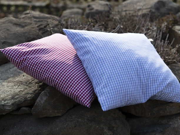 Barbara Raggl - Kissenmanufaktur Zwei karierte Kissen, eins rot-weiß, das andere blau-weiß, auf Steinen.