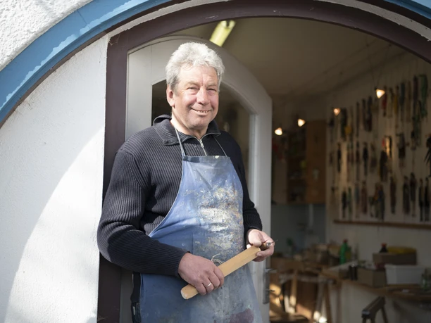 Oberammergauer Schnürlkasperl Ein Mann steht lächelnd in einer Tür, trägt eine Schürze und hält ein Holzstück.