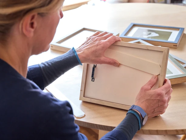 naïndesign Person prüft einen Holz-Bilderrahmen an einem Tisch mit anderen Rahmen.