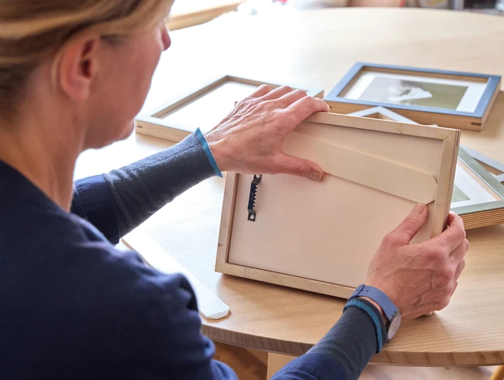 naïndesign Person prüft einen Holz-Bilderrahmen an einem Tisch mit anderen Rahmen.