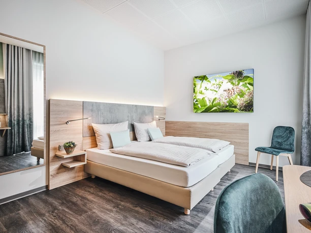 Helles Hotelzimmer mit Doppelbett, Pflanzenbild an der Wand, schlichter Dekor und Holzboden.