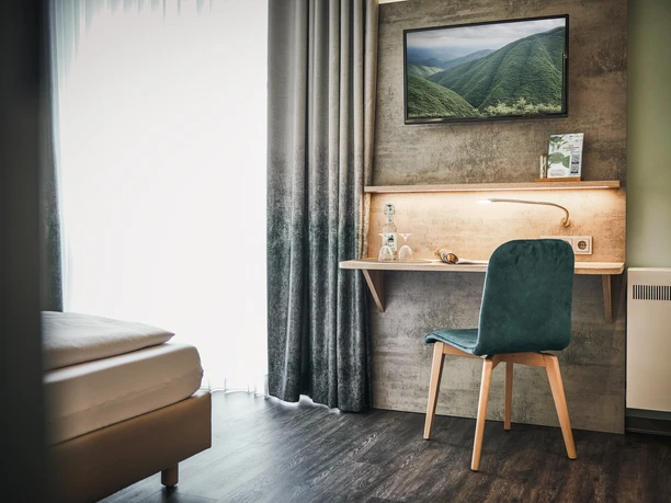 Modernes Hotelzimmer mit Schreibtisch, grünem Stuhl und Naturbild an der Wand. Helle Einrichtung.