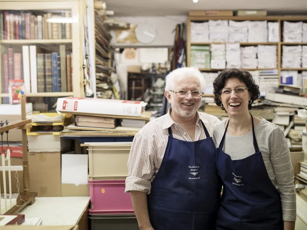 Buchbinderei Matheisl Zwei Personen in einer Buchbinderei, umgeben von zahlreichen Büchern und Materialien.