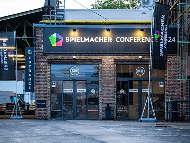 Spielmacher Conference draußen Spielmacher-Konferenzgebäude mit beleuchtetem Eingang, Industriefassade und Fahnen bei bewölktem Himmel.