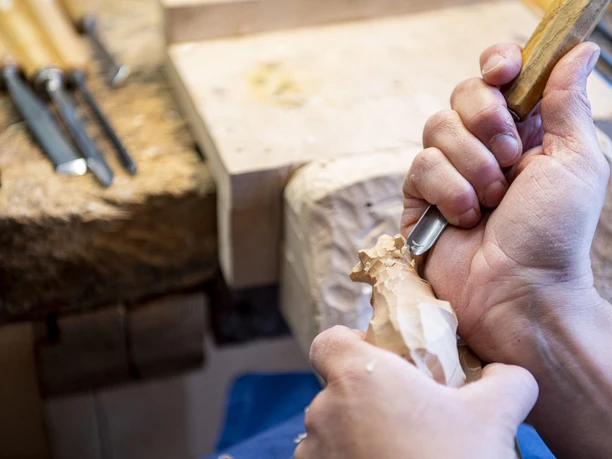 Marion Jochner Hände schnitzen mit einem scharfen Werkzeug an einem Holzstück, Werkzeuge im Hintergrund.