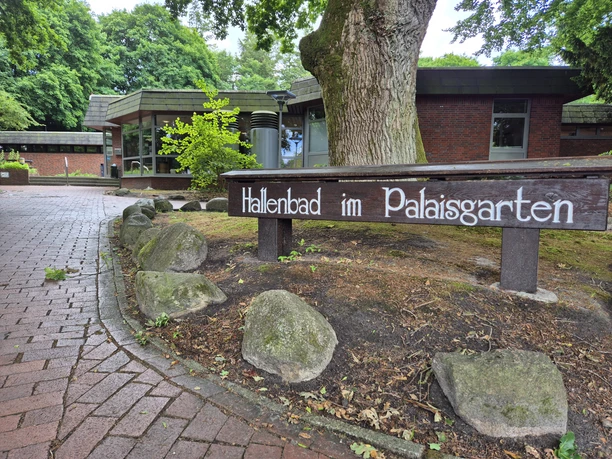 Aussenansicht Hallenbad Ein gepflegter Eingang zu einem Hallenbad im Palaisgarten, umgeben von Bäumen und einem Weg aus rötlichen Steinen.