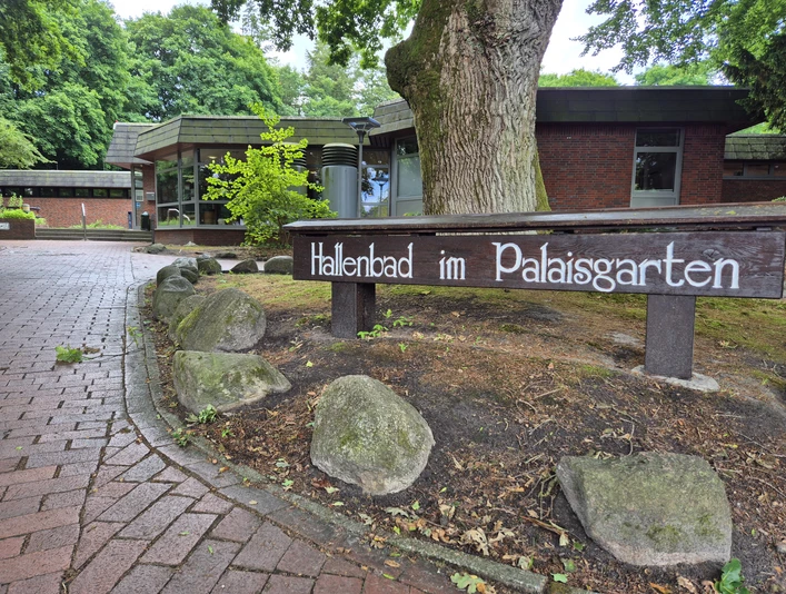 Aussenansicht Hallenbad Ein gepflegter Eingang zu einem Hallenbad im Palaisgarten, umgeben von Bäumen und einem Weg aus rötlichen Steinen.