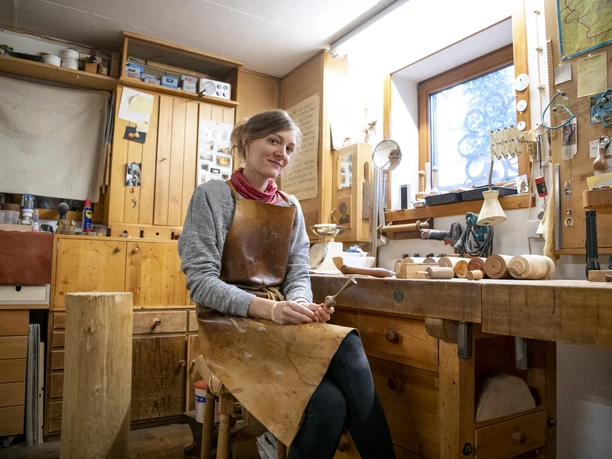 Holzdesign Eva Engler In einer Holzwerkstatt sitzt eine lächelnde Person an einem Arbeitstisch.
