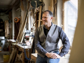 Wohnladen & Schreinerei Geiger Mann in Schreinerwerkstatt sitzt entspannt auf einem Arbeitstisch, umgeben von Werkzeugen.
