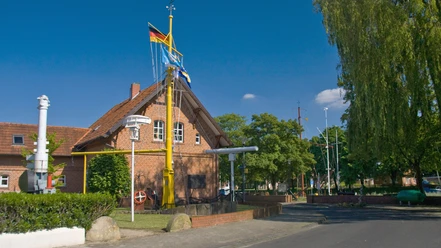2025-Schifffahrtsmuseum_Haren_-Schleusenwaerterhaus-Bild1.jpg Backsteingebäude mit Schiffsmodellen, Masten und Flaggen unter blauem Himmel in Haren an der Ems.