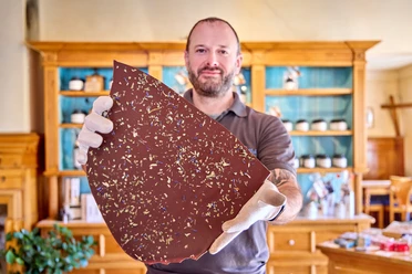 Chocowerk Wallgau Mann hält große Schokoladentafel mit Blütenstreuseln in einer Ladenumgebung.