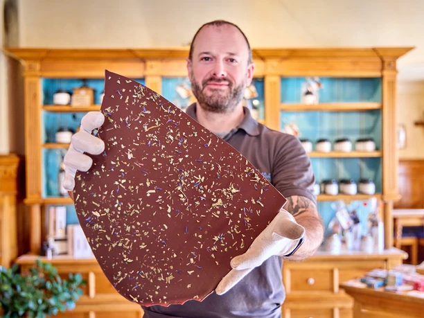 Chocowerk Wallgau Mann hält große Schokoladentafel mit Blütenstreuseln in einer Ladenumgebung.
