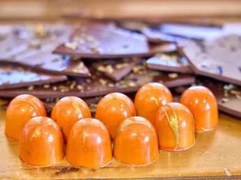 Chocowerk Wallgau Orange Pralinen auf Holztisch, dahinter Schokoladenstücke mit Blüten.